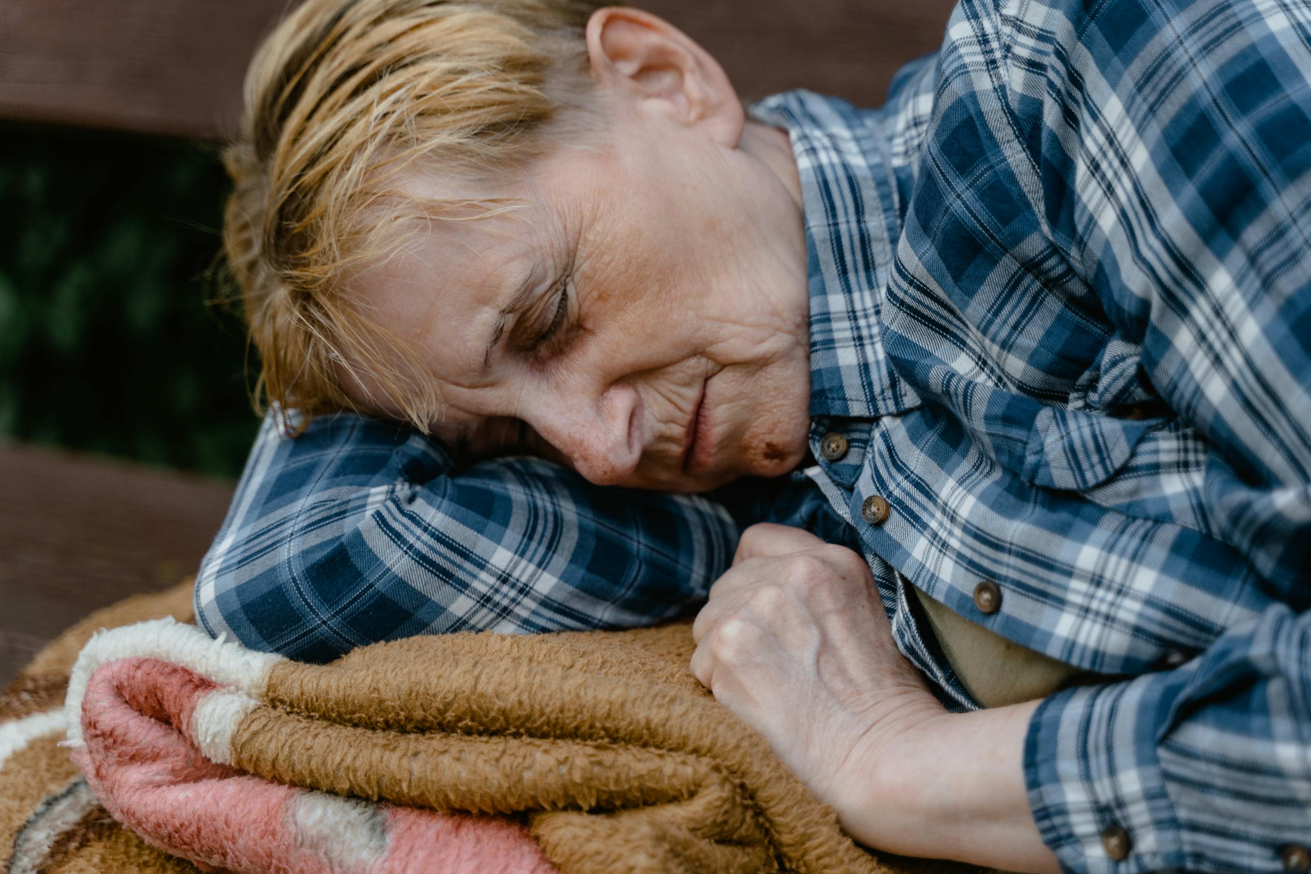 Close-up of a serene elderly woman sleeping outdoors on a bench, wrapped in a blanket.